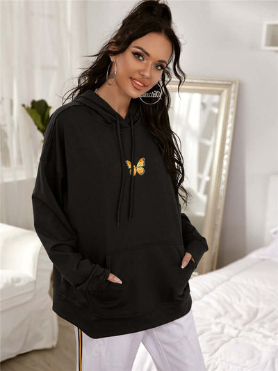 Woman wearing a black printed drop-shoulder hooded sweatshirt with butterfly design, styled comfortably in a cozy room.