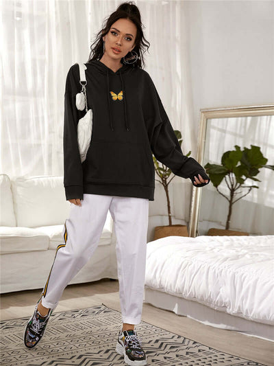 Woman wearing a printed drop-shoulder hooded sweatshirt with butterfly design, paired with white pants and sneakers.