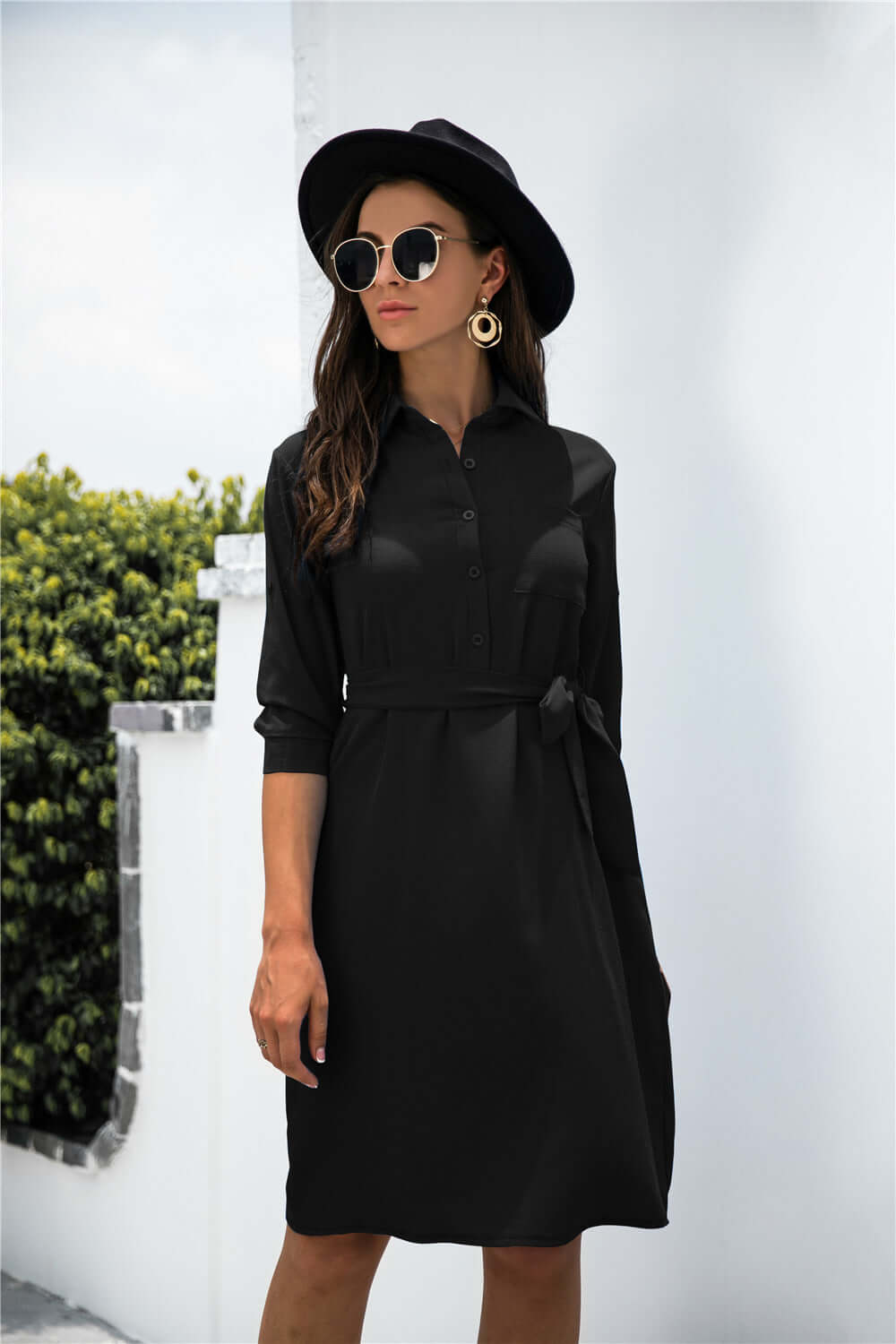 Solid color button lapel mid-sleeve shirt dress in black, styled with sunglasses and hat against a greenery backdrop.